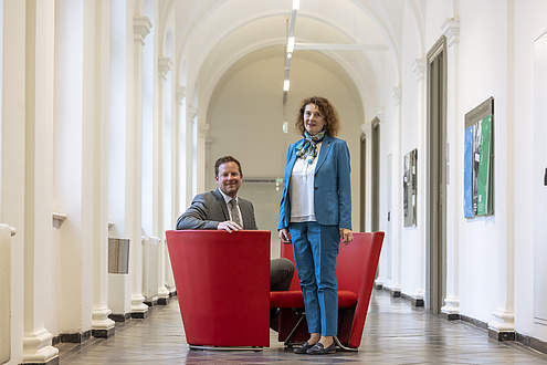 A woman and a man smile at the camera. The woman is standing and the man is sitting on a red armchair to her left.