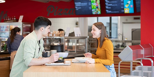 Ein junger Mann und eine junge Frau mit Tellern vor sich sitzen sich an einem Tisch in einer Kantine gegenüber.
