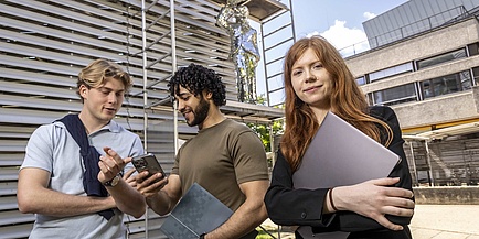 Zwei Studenten unterhalten sich am Campusgelände, daneben steht selbstbewusst eine Studentin.